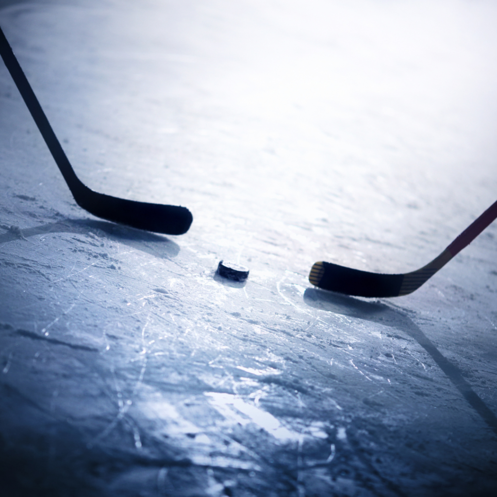 Two hockey sticks with a puck between them on the ice.